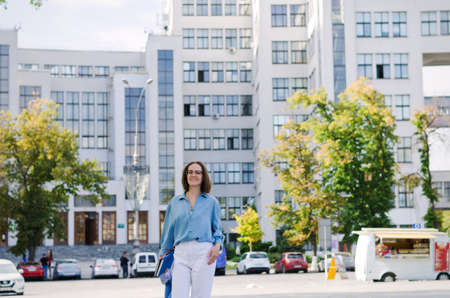 Urban portrait of young elegant business woman in eye glasses and casual clothes in city. Woman walking in green park, relaxing, smiling in sunny day. Hobby and leisure.の写真素材