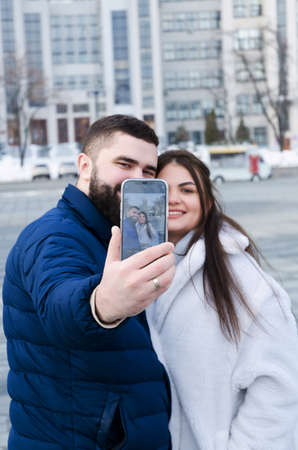 Love story of couple walking in city. Young business man in a blue jacket with a beard. And a cute woman in long coat. talking on the phone, making a selfie. Bloggers. Family holiday, dateの写真素材
