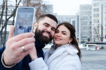 Love story of couple walking in city. Young business man in a blue jacket with a beard. And a cute woman in long coat. talking on the phone, making a selfie. Bloggers. Family holiday, dateの写真素材