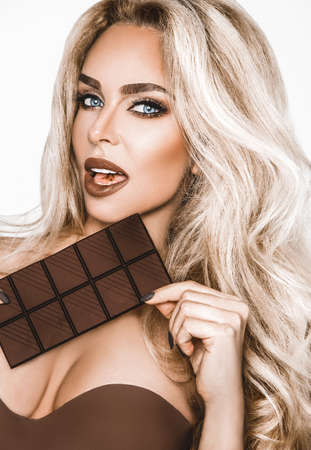 Portrait of a satisfied pretty girl biting chocolate bar and looking at camera isolated over white...