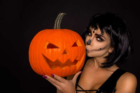 Sexy brunette woman in Halloween makeup and lingerie hold a pumpkin on a black background in the studio. Make-up skeleton, monster and witchの写真素材