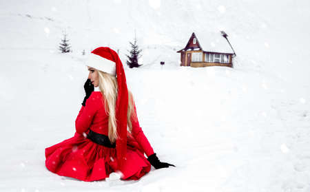 Merry Christmas. Beautiful sexy blonde woman in red dress and Santa clause hat on a background of mountains and snow. Beautiful, sensual girl for Christmas. Christmas concept.の写真素材