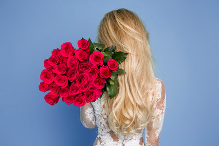 Beautiful woman holds bouquet with red roses. Vogue fashion style studio portrait of sexy girl in white elegant dress on blue backgroundの写真素材