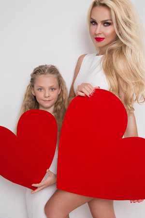 Beautiful blonde woman mother and daughter standing on a white background and holds a red heart. Lovely family. Valentines day and mothers day - concept.の写真素材