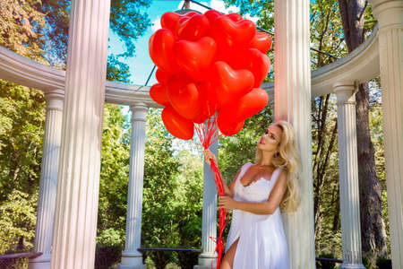 Bride in a wedding dress is walking in a beautiful garden and holding red balloons. Set of Air Balloons. Wedding fashion. Valentines day and bride at the wedding party.の写真素材