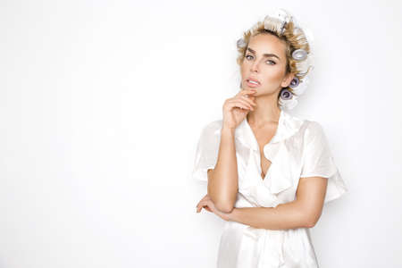 Pretty woman in hair curlers dressed in bathrobe isolated on white background. Sexy woman preparing...