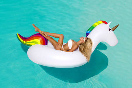 Beautiful sexy blonde model in a elegant bikini with a inflatable white unicorn in pool. Hit the summer. Girl with trendy, colorful blown unicorn mattress. Summertime. Bikini fashion.の写真素材