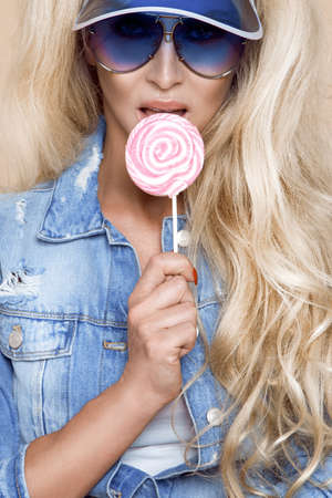 Sexy girl eating lollipop. Beauty glamor model woman with trendy jeans jacket, sunglasses and beautiful makeup holding pink sweet colorful lollipop candy, on beige, pastel background. Summertime.の写真素材