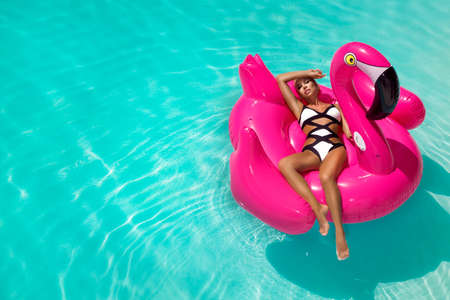Beautiful sexy tanned woman in a swimming pool sitting on an inflatable pink flamingo in elegant...