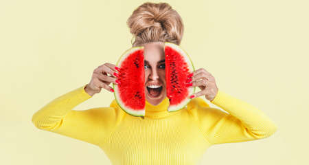 Happy young woman in colored dress holding slice of a watermelon isolated over yellow background....