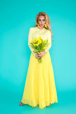 Spring fashion Woman. Beautiful happy woman with colorful dress, holding a bouquet of spring flowers on blue background in studio. Fashion photo. Spring flowers - concept. Woman with Easter tulips.の写真素材