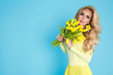 Spring fashion Woman. Beautiful happy woman with colorful dress, holding a bouquet of spring flowers on blue background in studio. Fashion photo. Spring flowers - concept. Woman with Easter tulips.の写真素材