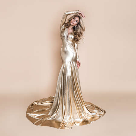 Elegant fashion. Stunning blonde woman in elegant long gold dress and perfect hairstyle on beige background. Luxury evening fashion. Glamor fashion model. Elegance. Golden dress.の写真素材