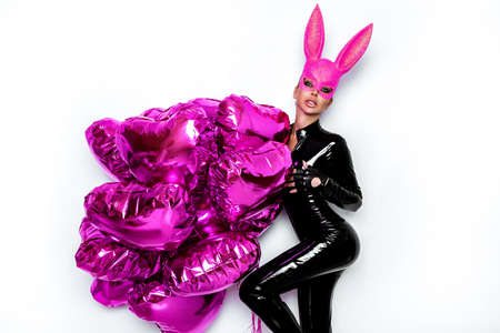 Sexy blonde woman posing in latex black costume and pink bunny mask and pink balloons on white background. Latex lingerie. Naughty girl. Halloween costume. Woman with balloons.の写真素材