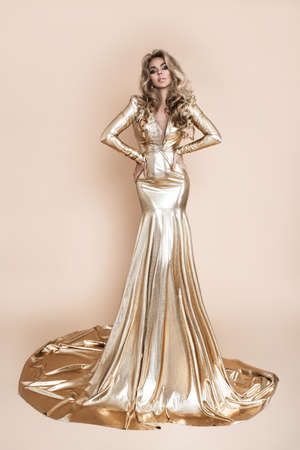 Elegant fashion. Stunning blonde woman in elegant long gold dress and perfect hairstyle on beige background. Luxury evening fashion. Glamor fashion model. Elegance. Golden dress.の写真素材