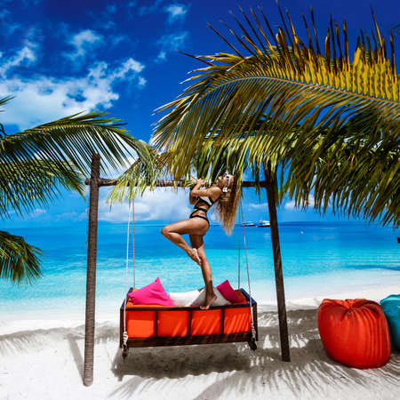 Beautiful sexy tanned woman in elegant bikini on Maldives island. Young glamor girl is posing on swing in paradise. Perfect body bikini model on Maldives beach. Bikini fashion. Luxury travel.の写真素材