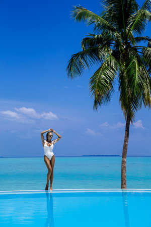 Elegant tanned woman in white swimsuit in pool on tropical Maldives island. Beautiful bikini body girl in pool with view on horizon. Sexy model near the pool on beautiful Indian ocean landscape. Travel. Maldives landscape.の写真素材