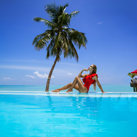 Elegant tanned woman in red swimsuit in pool on tropical Maldives island. Beautiful bikini girl in pool with view on horizon. Sexy model near the pool on beautiful Indian ocean landscape. Travel.の写真素材