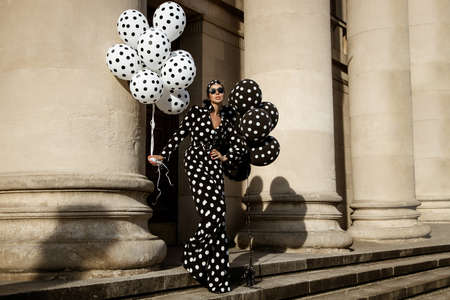 Spring, summer fashion. Glamor, stylish elegant woman in polka dot jumpsuit is holding balloons with dots. Fashion model in outfit with polka dots in the city. 60's style. Retro fashionの写真素材