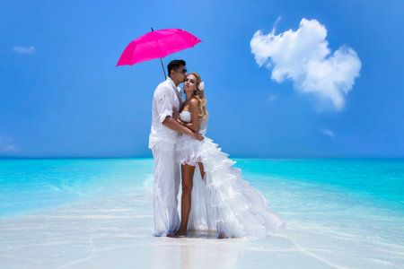 summer love. Beautiful happy young couple in wedding clothes and pink umbrella is standing on a beach in the Maldives. Engagements and wedding on the beach on the paradise island of Maldives.の写真素材