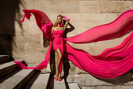 Glamour, stylish elegant woman in pink fluttering dress is posing outdoor on a summer day. Female model in amazing long dress. Vogue. Couture.の写真素材