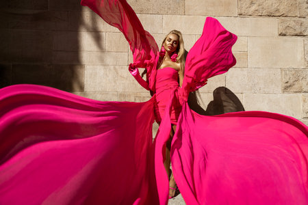 Glamour, stylish elegant woman in pink fluttering dress is posing outdoor on a summer day. Female model in amazing long dress. Vogue. Couture.の写真素材