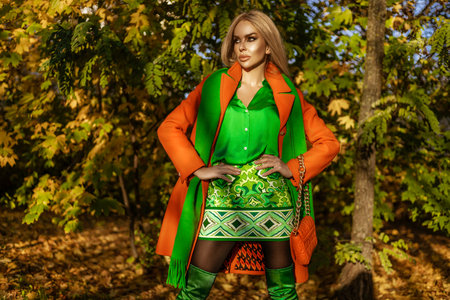 Beautiful woman wearing a orange coat and elegant green dress is posing outdoors on a sunny autumn day.の写真素材