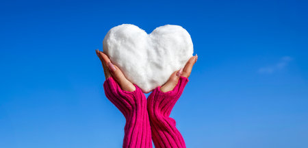 Heart-shaped snow held by hands in cozy pink sleeves against a vibrant blue sky, symbolizing winter love, romance, and warmth in a minimalist and artistic seasonal composition.の写真素材