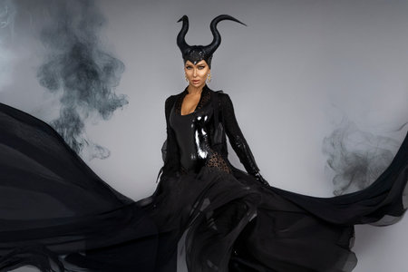 Dark queen with black horns and red lips in dramatic Halloween portrait. Witch with smoke —...