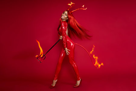Halloween devil woman in red latex costume with fire horns, tail and trident posing on red...