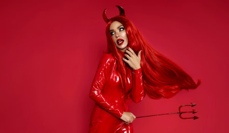 Halloween devil woman in red latex costume with fire horns, tail and trident posing on red...