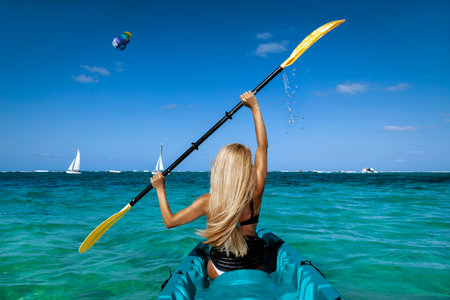 Woman kayaking in turquoise ocean water on Mauritius, holding paddle above head with splashing drops under clear blue sky, adventure travel and tropical lifestyle conceptの写真素材
