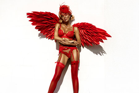 Seductive Blonde Woman in Red Lingerie with Angel Wings Posing Against White Wall in Bright Sunlight