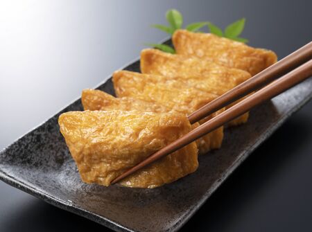 Japanese Inari Sushi on a black plate set against a black background is lifted with chopsticksの写真素材