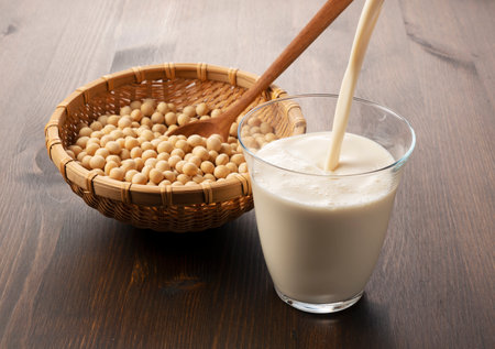 Pouring soy milk into a glass placed on a wooden background. Soybeans in a bamboo colander in the backgroundの写真素材