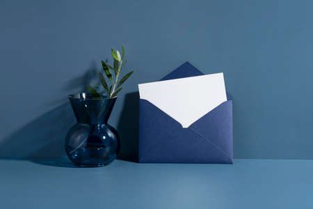 Combination of glass vase and olive branches. Sunlight table background. White card in a dark blue seal leaning against a blue wall. Invitation mockup, long shadow.の写真素材