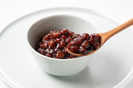 Boiled azuki beans in a small bowl placed against a white background. Scoop them up with a wooden spoon.の写真素材