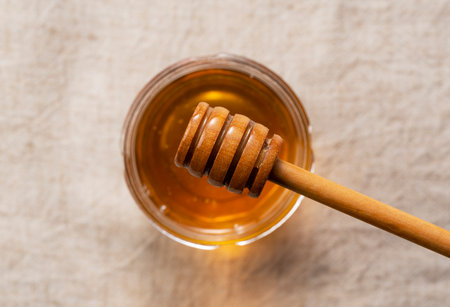 Honey in glass container and wooden honey dipper on cloth background. Honey drips from the honey dipper. View from above.の写真素材