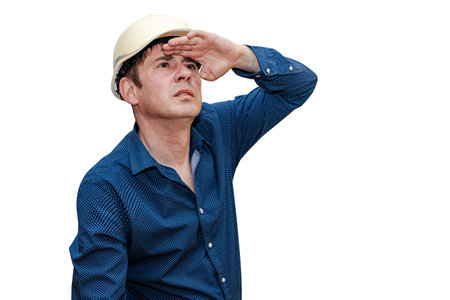 A man in a construction helmet on a white isolated background looks into the distance with his hand to his eyesの写真素材
