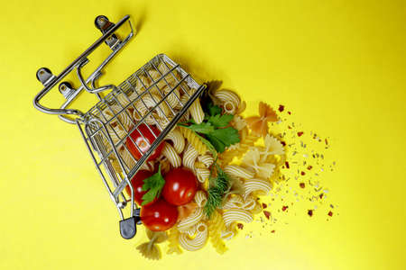 Inverted cart from the store with different types of pasta, ripe cherry tomatoes, spices and fresh herbs. Bright yellow background. Products for cooking delicious food. Healthy eatingの写真素材