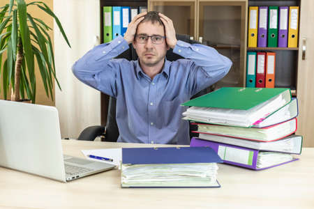 Boss businessman at his desk in the office with a pile of folders. Holds his head with his hands....