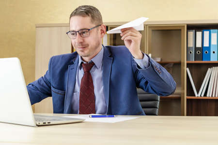 Distracted from computer work happy 30s young bearded caucasian man in glasses throwing paper airplane, enjoying break pause time alone in modern office, entertaining having fun relaxing indoorsの写真素材