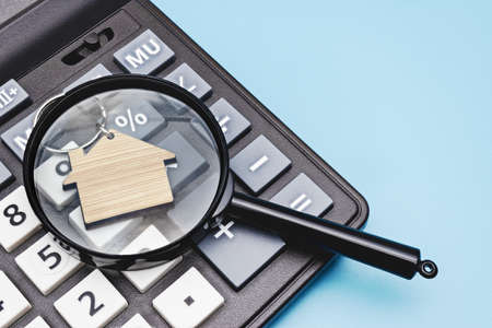 The concept of mortgage, sale and rental of housing and real estate. Mortgage credit lending. Calculator with a wooden house under a magnifying glass on a blue background. Copy spaceの写真素材