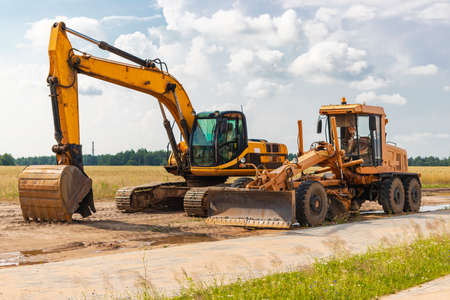 The excavator and the grader stand side by side against the blue sky. Heavy construction earthmoving...