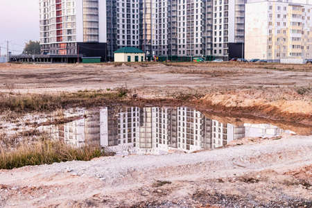 A large puddle after rain at the construction site of a large residential facility. Reflection in the water of the construction site and cranes against the background of the sunset skyの写真素材