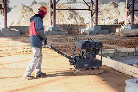 A worker compacts the earth with a vibrating rammer on a construction site on a bright sunny day....