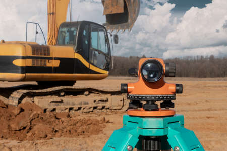 Level with an excavator in the background. Construction level or theodolite. Geodetic instruments and equipment for the construction and earthworksの写真素材