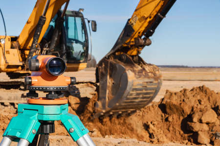 Level with an excavator in the background. Construction level or theodolite. Geodetic instruments and equipment for the construction and earthworksの写真素材