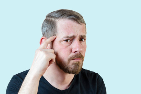Serious young bearded man touches his forehead with his finger. Focuses attention on the problem. Isolated portrait on a pastel backgroundの写真素材