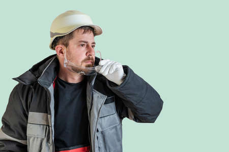 Positive friendly bearded male builder in overalls and safety hardhat and goggles looking at the...